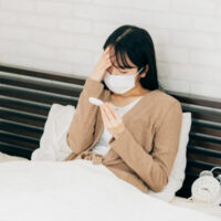 ベッドでマスクを着用し、体温計を見ながら額に手を当てる体調不良の女性