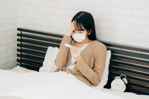 ベッドでマスクを着用し、体温計を見ながら額に手を当てる体調不良の女性